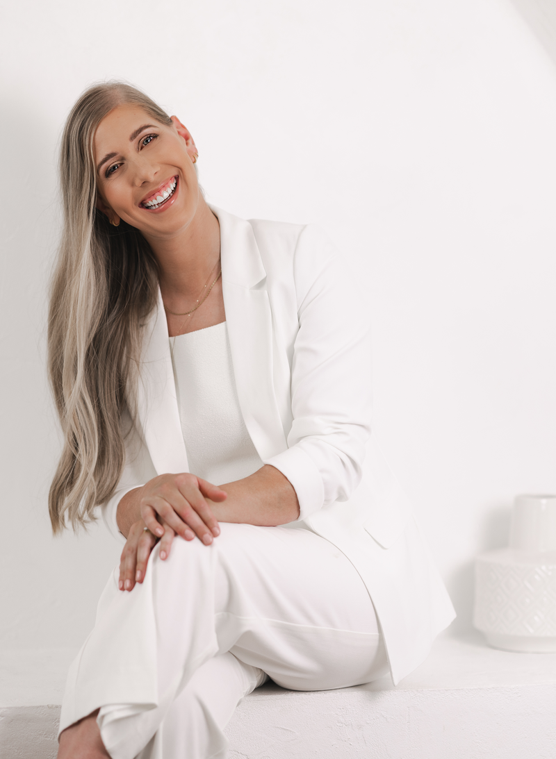Smiling woman seated in an all-white outfit, with long blonde hair, in a minimalist white space. She exudes confidence and warmth while wearing a white blazer and trousers.