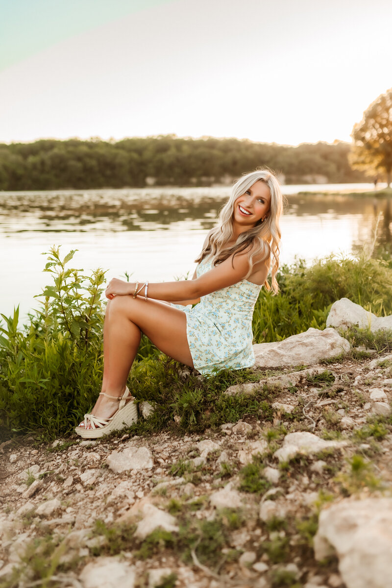 Beautiful senior girl smiles and sits on rock by lake in Kansas City with Senior photographers.