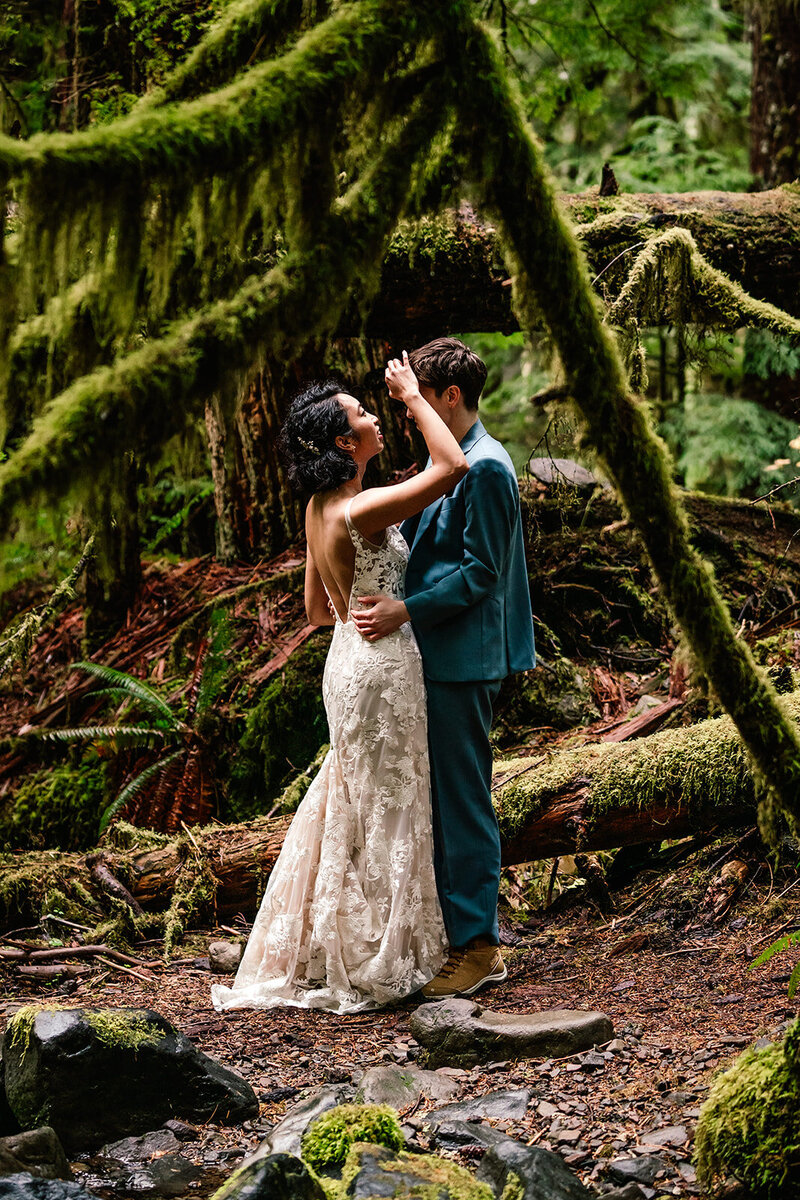a couple in wedding attire, stands deep in e forest. Their adventure elopement photographers shoot through the verdant hanging moss, framing the two brides with the greenery. 