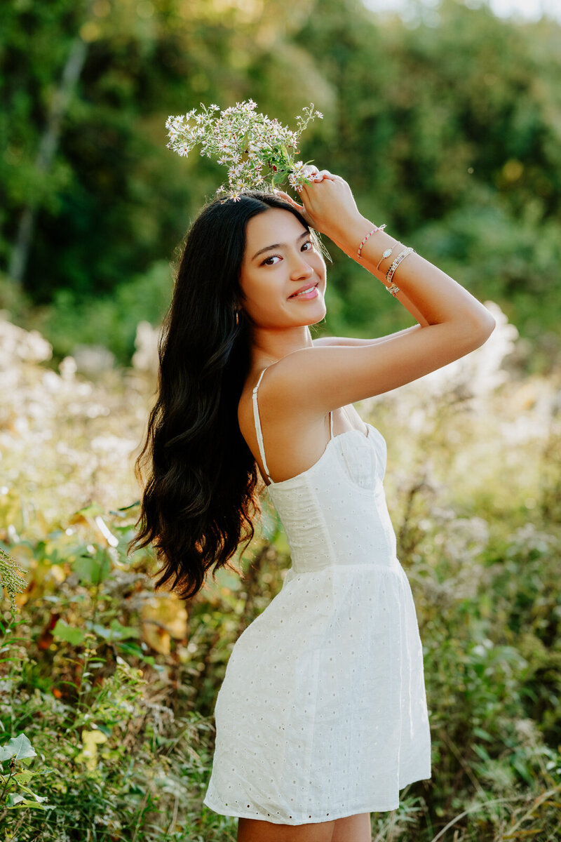 Girl wearing a white dress holding flowers over her head.
