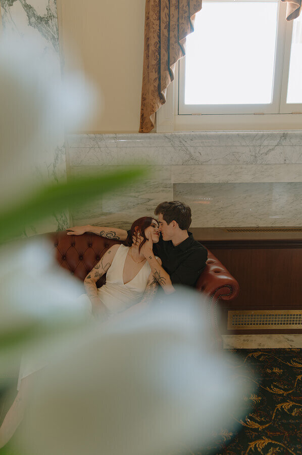 Sitting on a red leather couch inside of a nice government building, a man gently holds the face of his fiancé in his hand as he goes in to kiss her. 