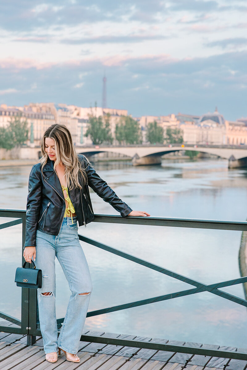 Stylish solo woman in a leather jacket and jeans, smiling confidently during a Paris photoshoot near the Louvre and Pont des Arts.
