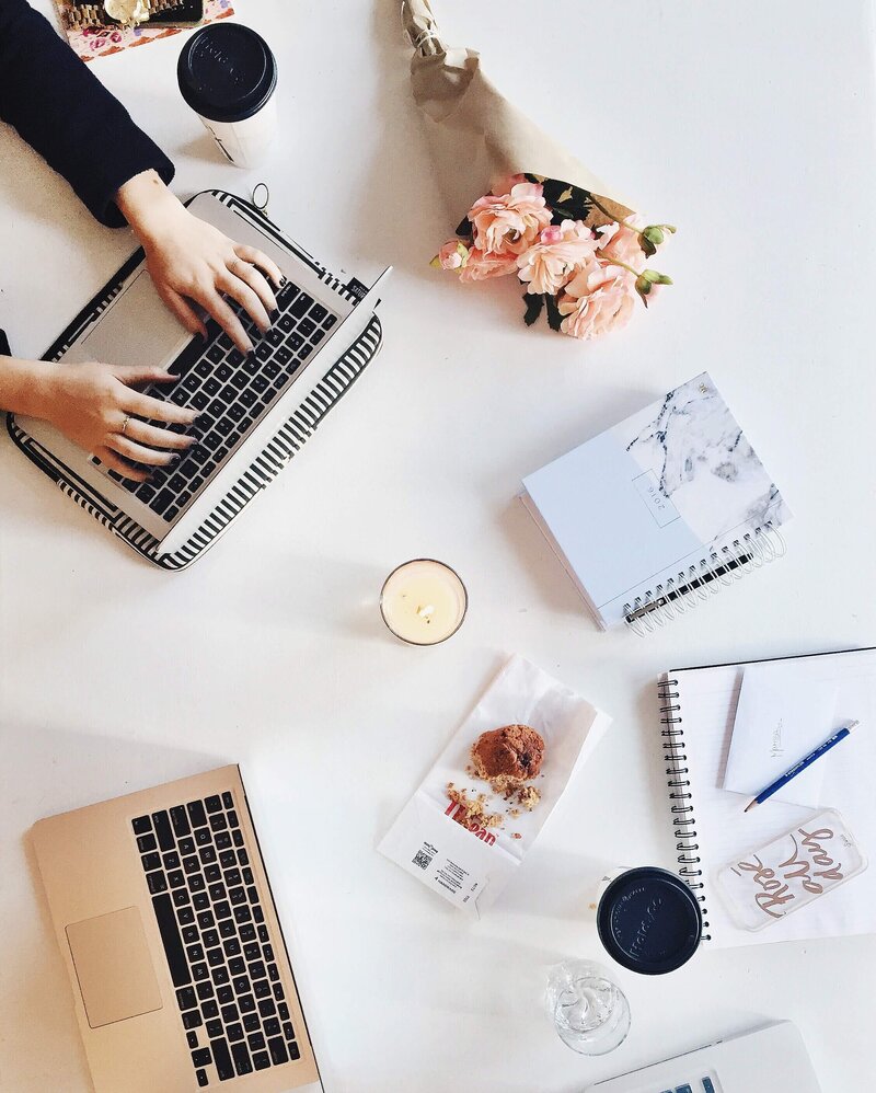 Flatlay of woman working on laptop laptop and other desk items like a bouquet of roses coffe candle bake good planner