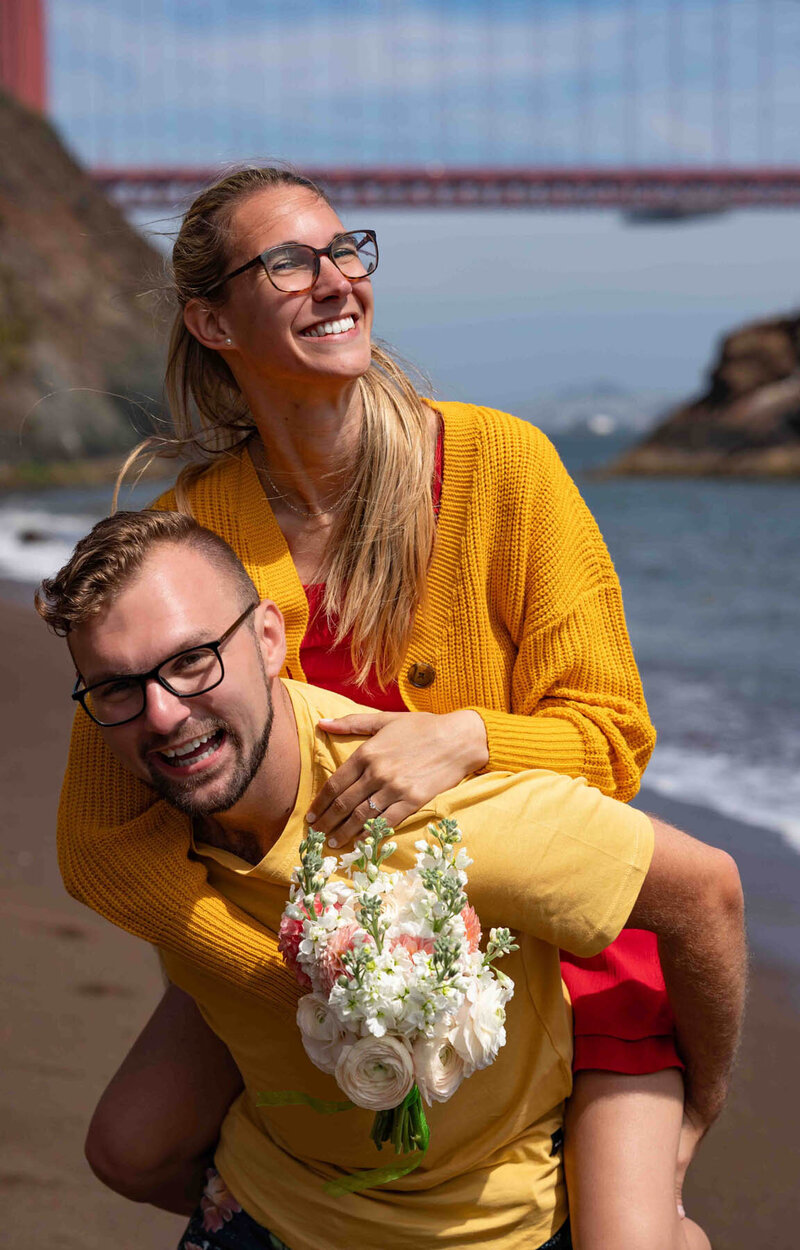 Smiling couple on piggyback at the beach following a surprise engagement with the golden gate bridge in the background