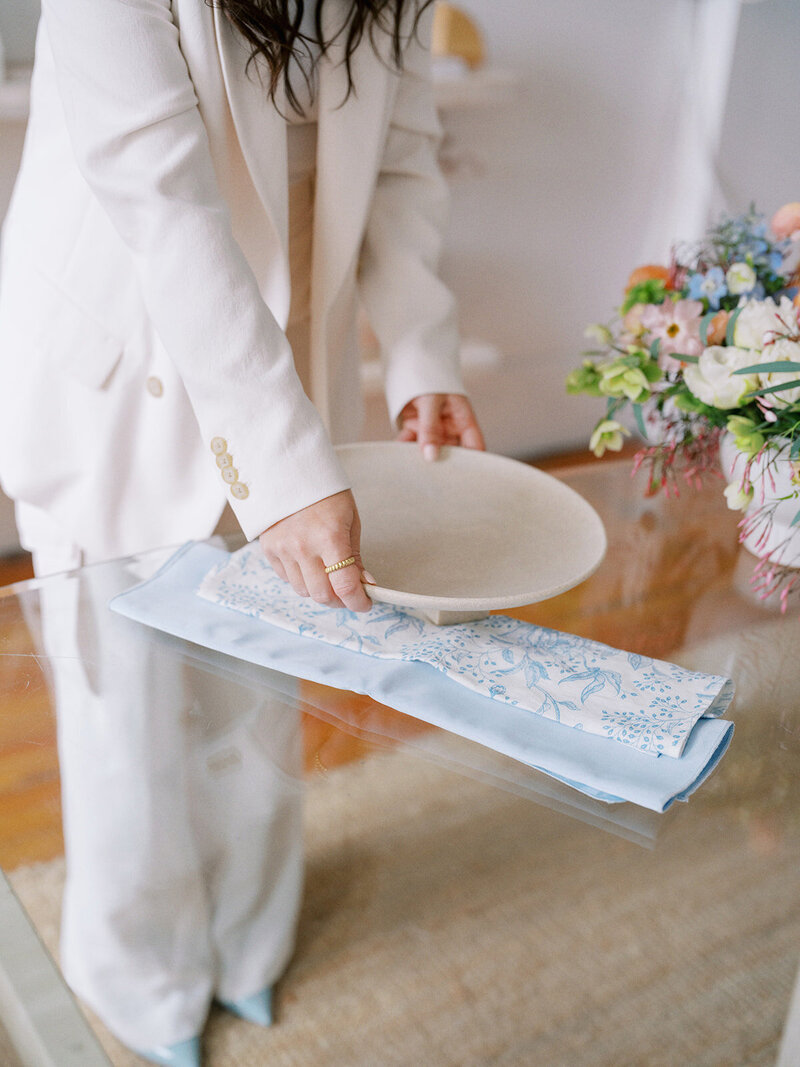 A person in a cream-colored suit sets a ceramic plate on a clear glass table next to a blue embroidered fabric and a floral arrangement, all perfectly styled for a luxury wedding planner website.