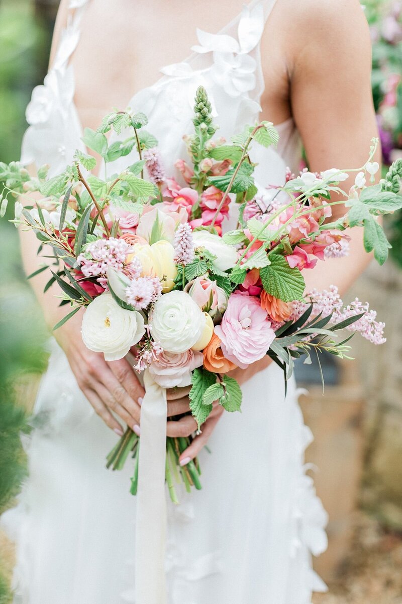 Bride holding a vibrant garden-style bouquet with ranunculus, roses, sweet peas, and seasonal greenery in soft pink, peach, and cream tones
