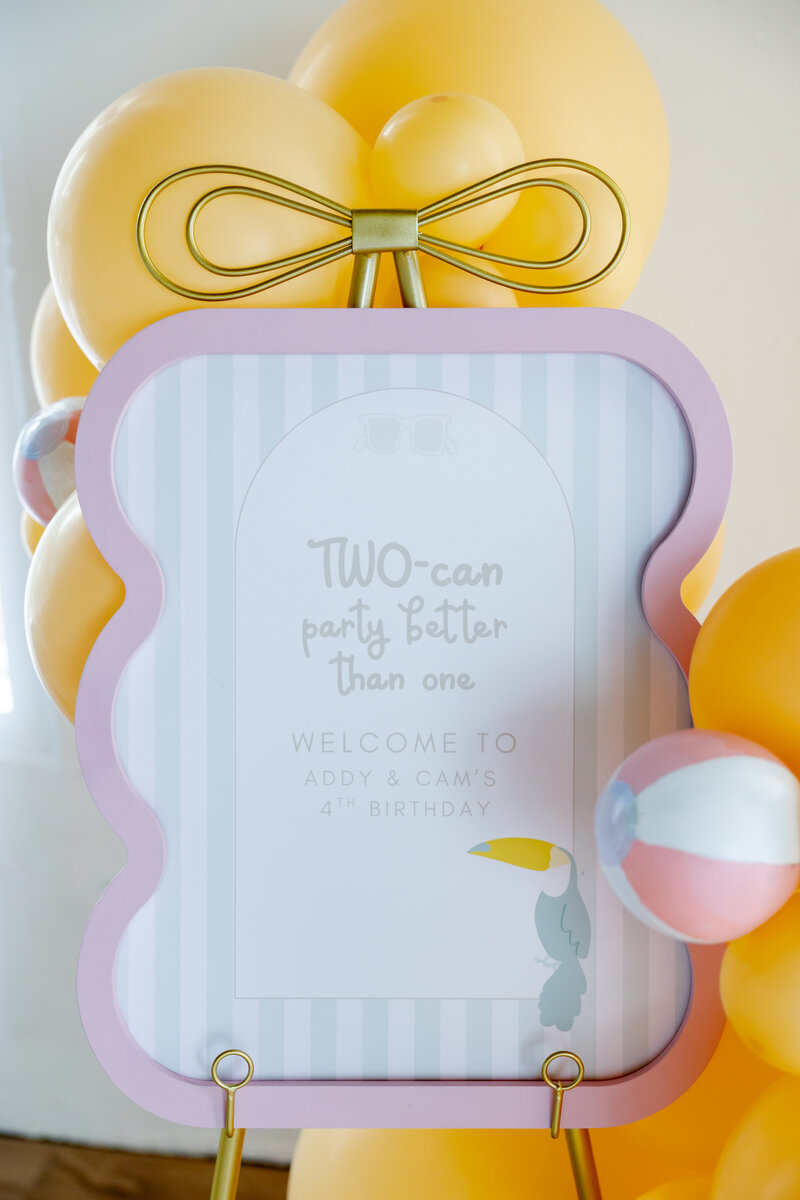 Pink and white sign on a gold easel with a peach balloon garland and small beach balls. 
