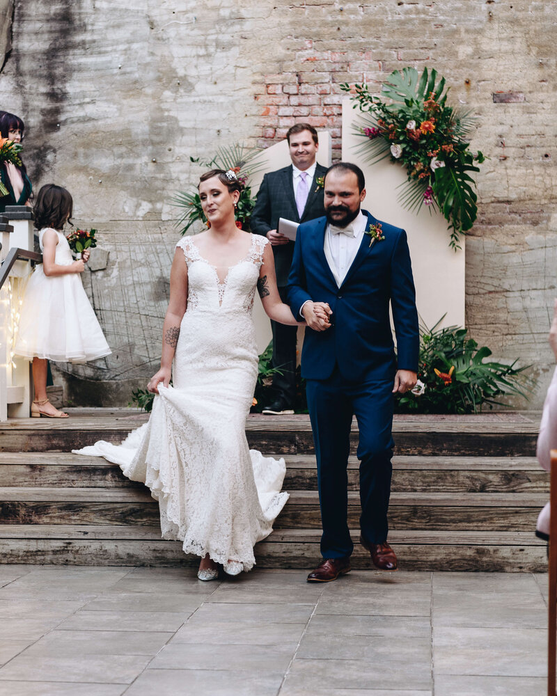 A bride in a white lace gown and a groom in a navy suit walk hand in hand down steps, smiling, after their wedding ceremony. A flower girl and officiant stand in the background near floral decorations.