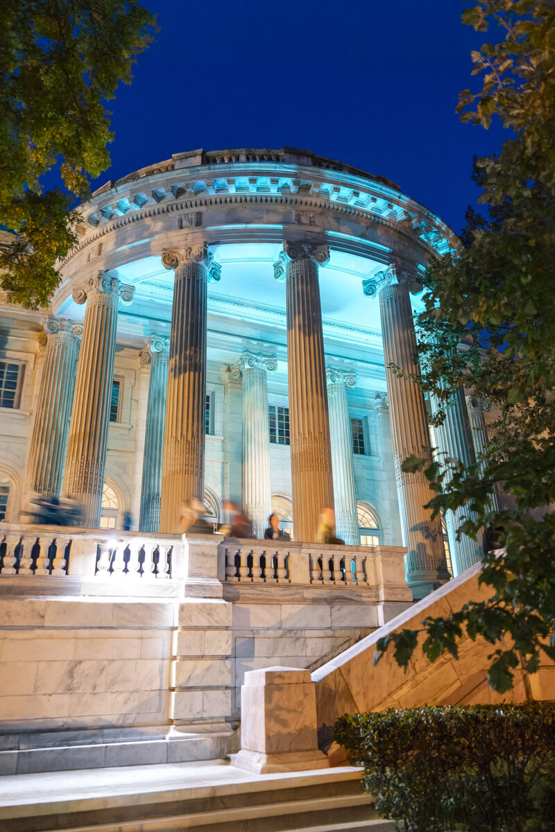 A neoclassical building with tall columns is illuminated with blue lights against a night sky. Stairs lead up to the entrance, and trees frame the scene on the left and right.