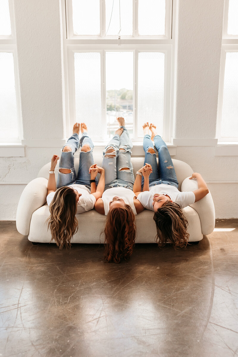 Three fitness instructors in white shirts and blue jeans lay on couch upside down during portrait studio session in Kansas City with Headshot Photographers.