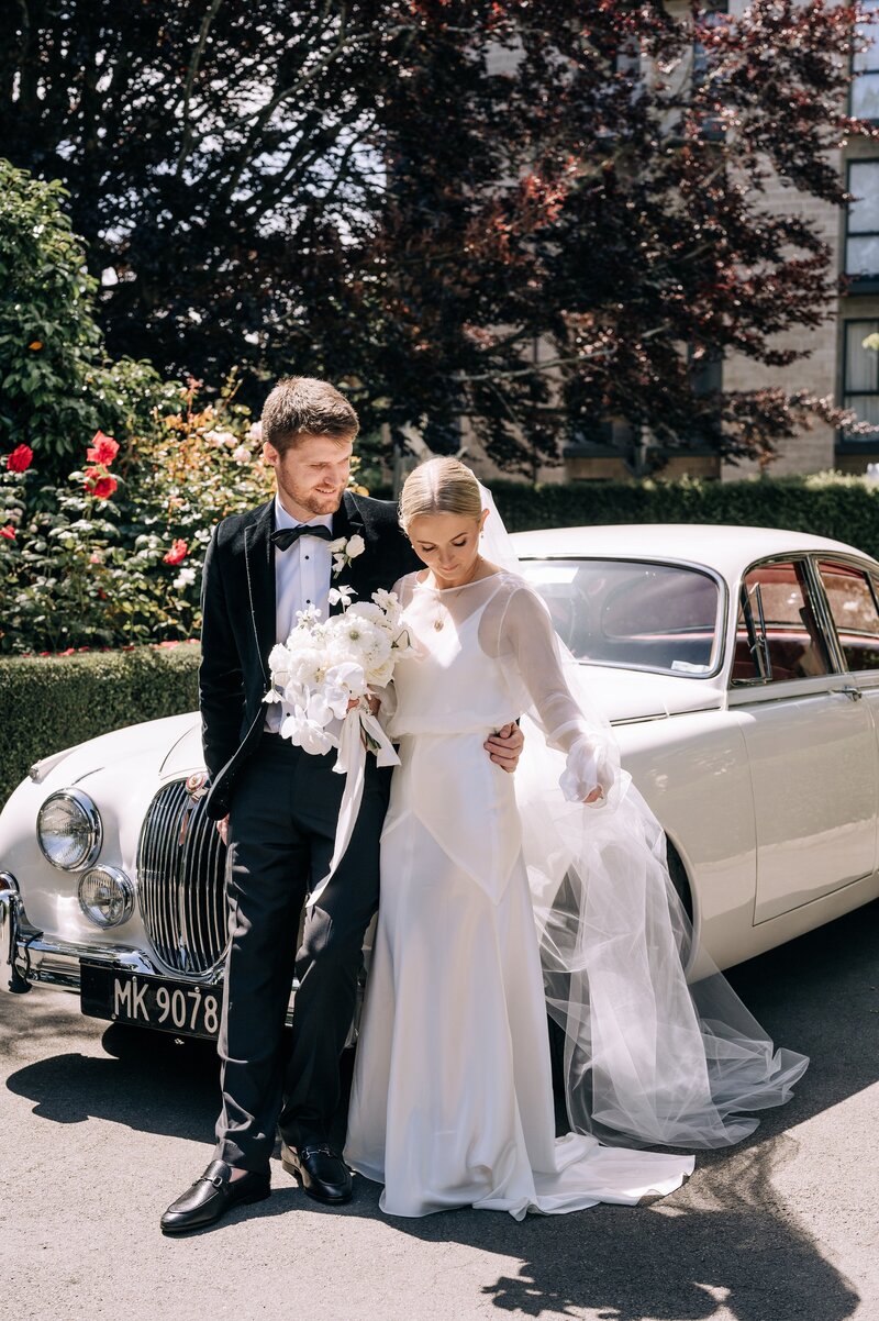 a bride in kenny and harlow dress with groom in front of a vintage white car at the rose historic chapel in christchurch