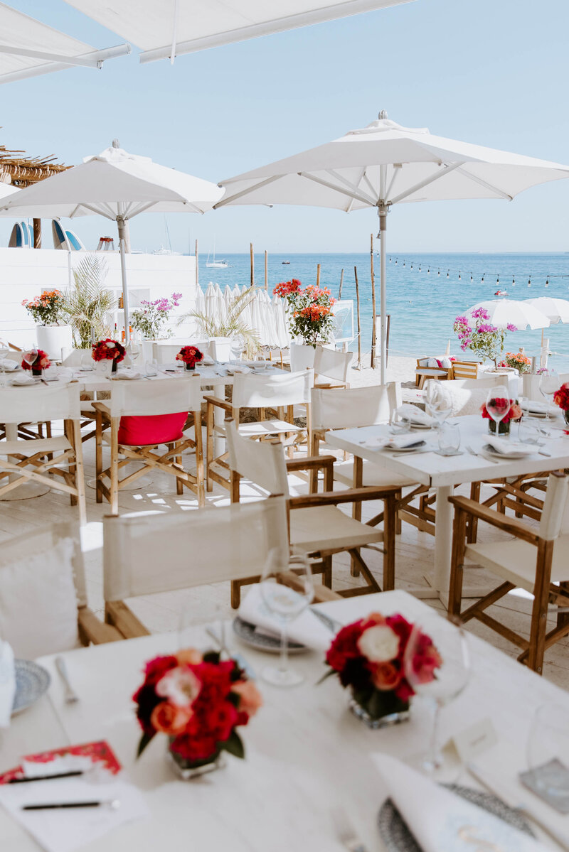 Outdoor wedding reception set up on an elevated patio on the beach with white tables, white umbrellas, and small red flowers