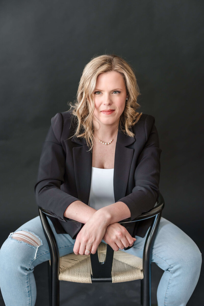 Modern headshot of Kelowna Naturopath wearing a black suit sitting in a modern chair for brand photoshoot.