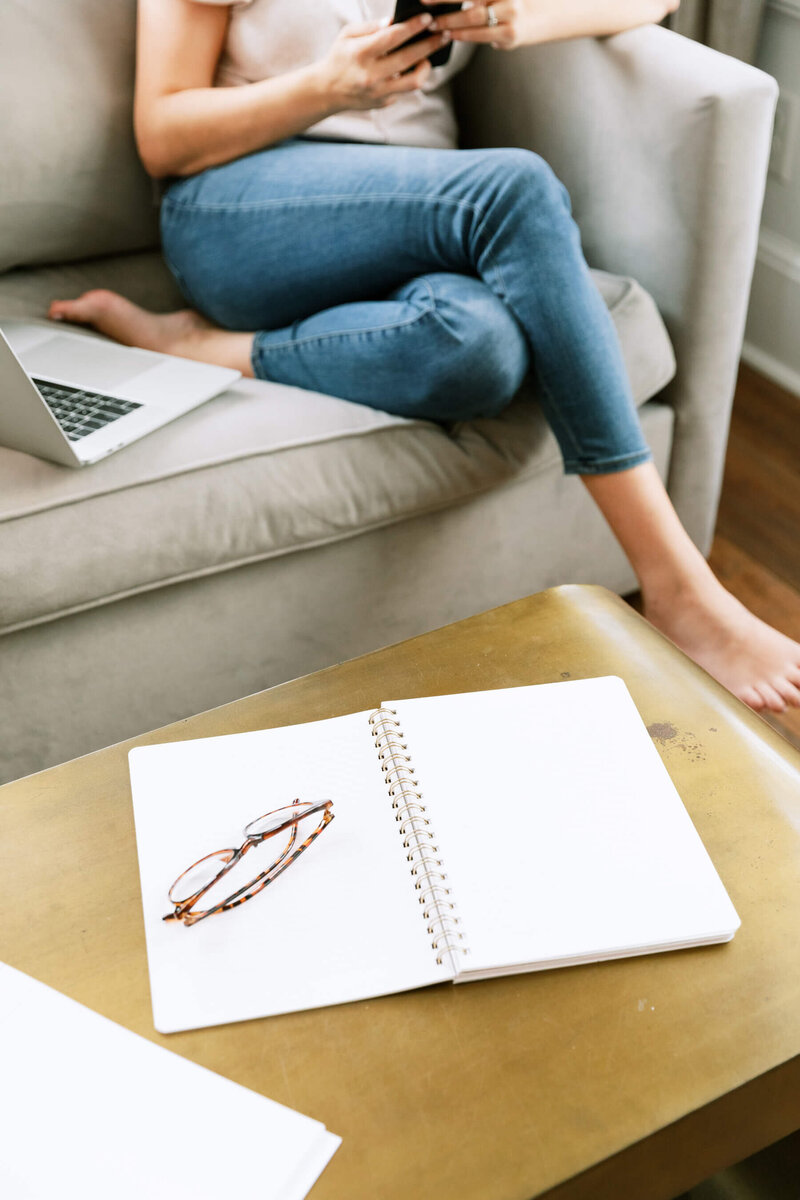 A woman relaxes on a couch, using her smartphone while her laptop rests beside her.