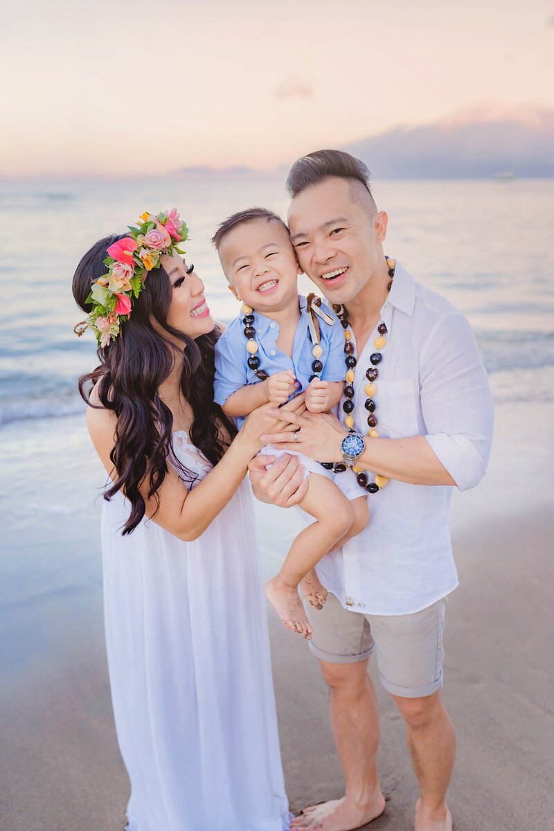 Child in blue shirt is held up by both parents as he smiles at the camera during their West Maui family photography session