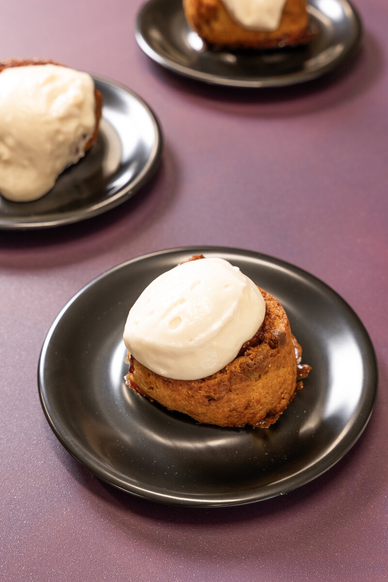 Close-up of a gluten-free cinnamon roll topped with cream cheese frosting on a dark plate, featured in Grain Artisan Bakery’s Thanksgiving pre-order menu in Snohomish, Washington.