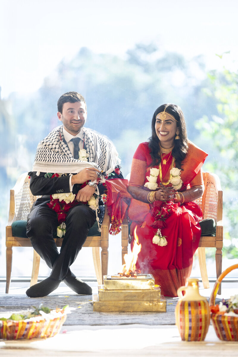 A couple sits side by side on chairs during a traditional Shaadi ceremony. The woman wears a red sari, and the man is dressed in a suit with a distinctive shawl. They are smiling and holding garlands, with a ceremonial fire burning in front of them.