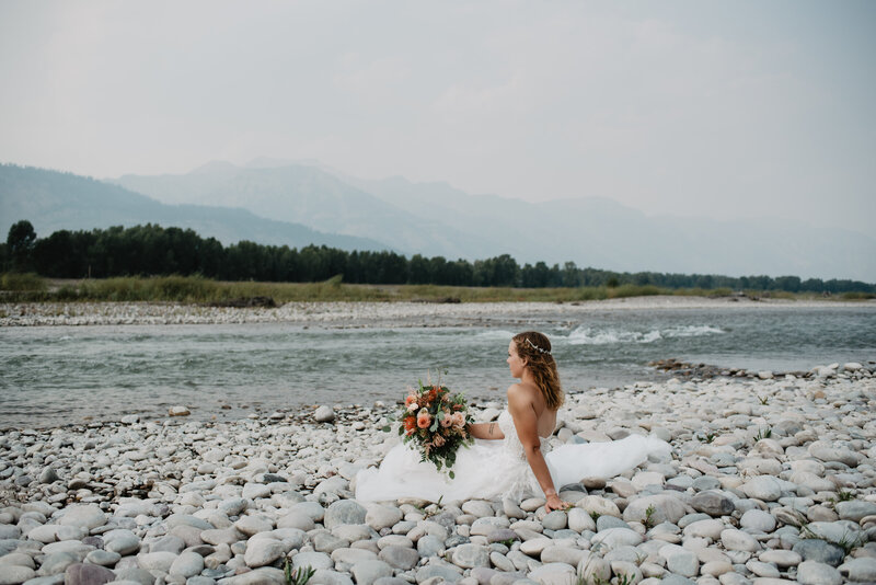 jackson hole photographers captures bride sitting on the bank of the river on the smooth river rocks while holding her bouquet on her lap as she leans backwards on her hand and watches the river flow