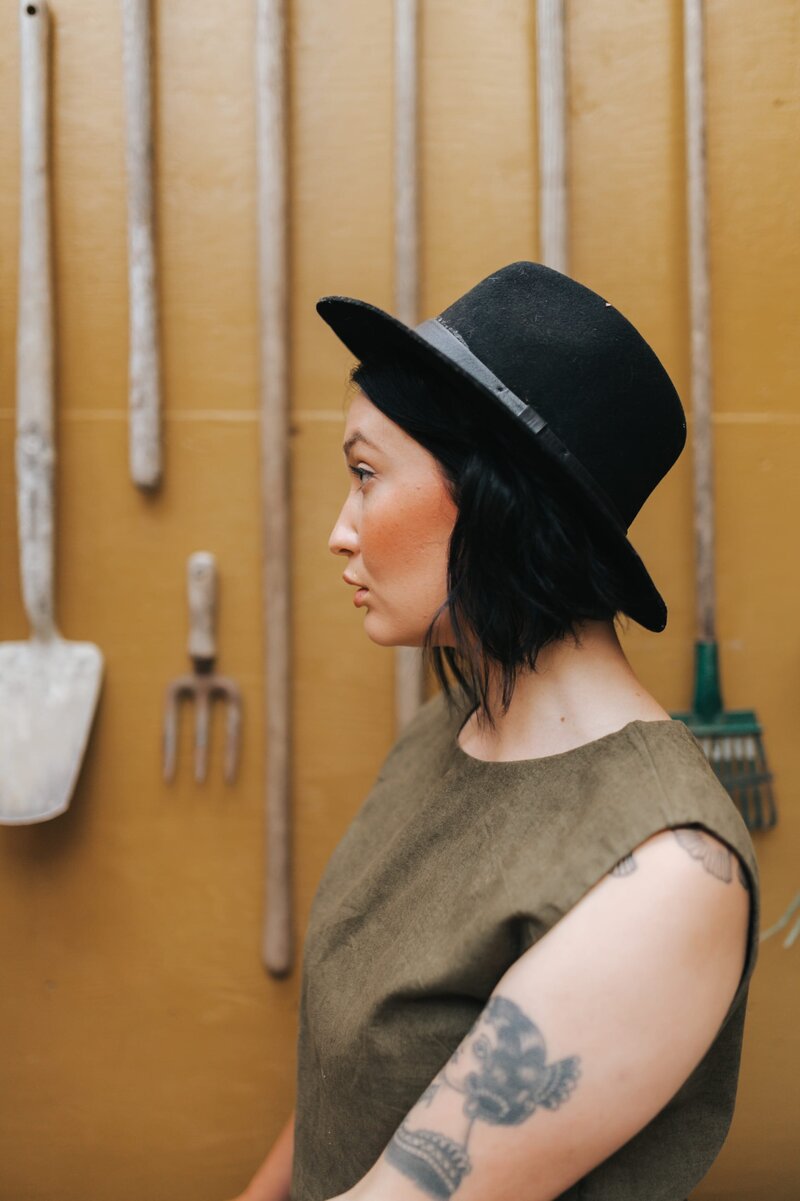 Woman with short, dark hair and a black hat faces the side with mustard-colored wall behind her and various hanging garden tools.