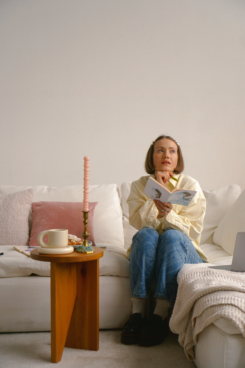 A person with short hairs sits ona. white couch in blue jeans, staring off into space as she dreams, a notebook and green pen in hand. beside her is a wooden coffee table with a cream mug and pink candle