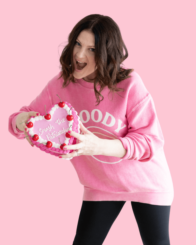 Emily, brand designer, in a pink sweatshirt that says "moody" is holding a heart shaped cake that says "smash the patriarchy."
