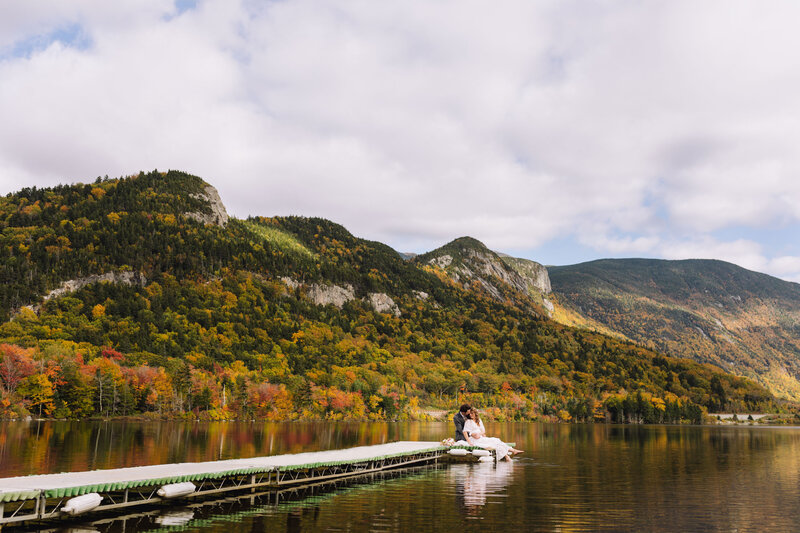 Man and Woman sit on the dock with the White Mountain foliage behind them