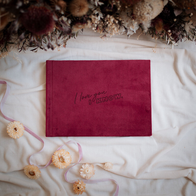 Burgundy velvet guest book with custom engraving on the cover in front of a dried flower arrangement