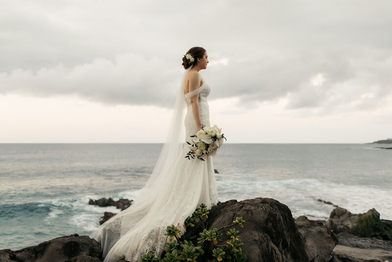 Bride stands on a rock over the ocean holding a white bouquet with a flower hair piece and veil