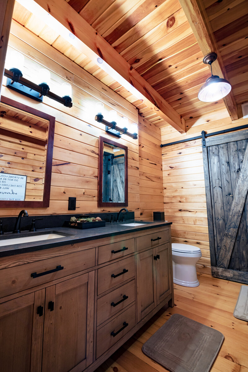 Interior view of a rustic bathroom with wooden walls and ceiling. Features include a vanity with dark cabinets, a vessel sink, and a sliding barn door. The space has a warm cabin aesthetic with wood throughout.