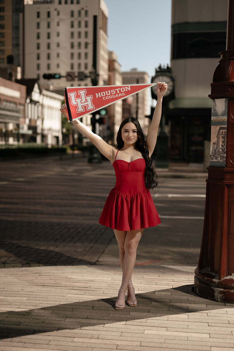 A woman in a red dress stands on a city street holding a red "Houston Cougars" pennant high above her head, exuding a cheerful and proud demeanor.