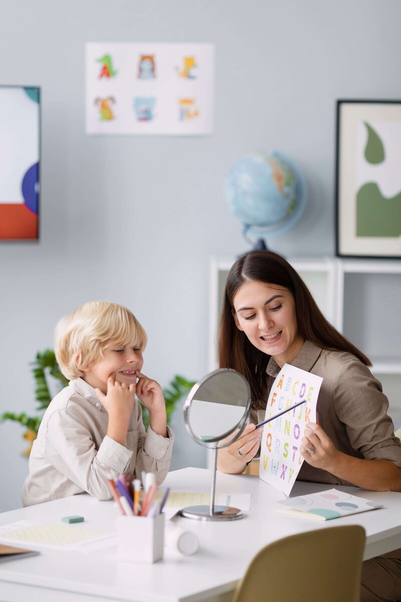 Speech-language pathologist using a mirror and letter chart to guide a young boy through articulation exercises during a speech therapy session in a bright, welcoming office.