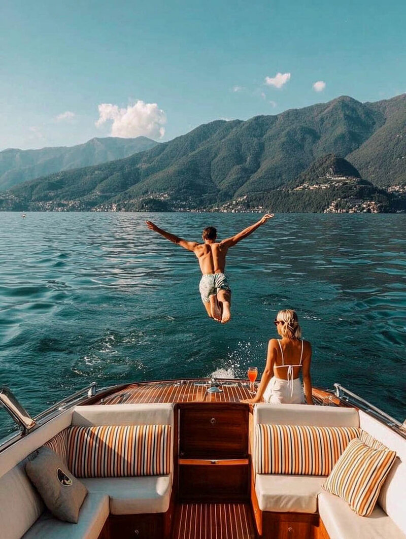 Couple enjoying a luxury European lake escape aboard a classic wooden speedboat, with the man diving into the water and the woman relaxing with a drink against a backdrop of lush mountains during custom itinerary. Tailored Oasis Travel Co.