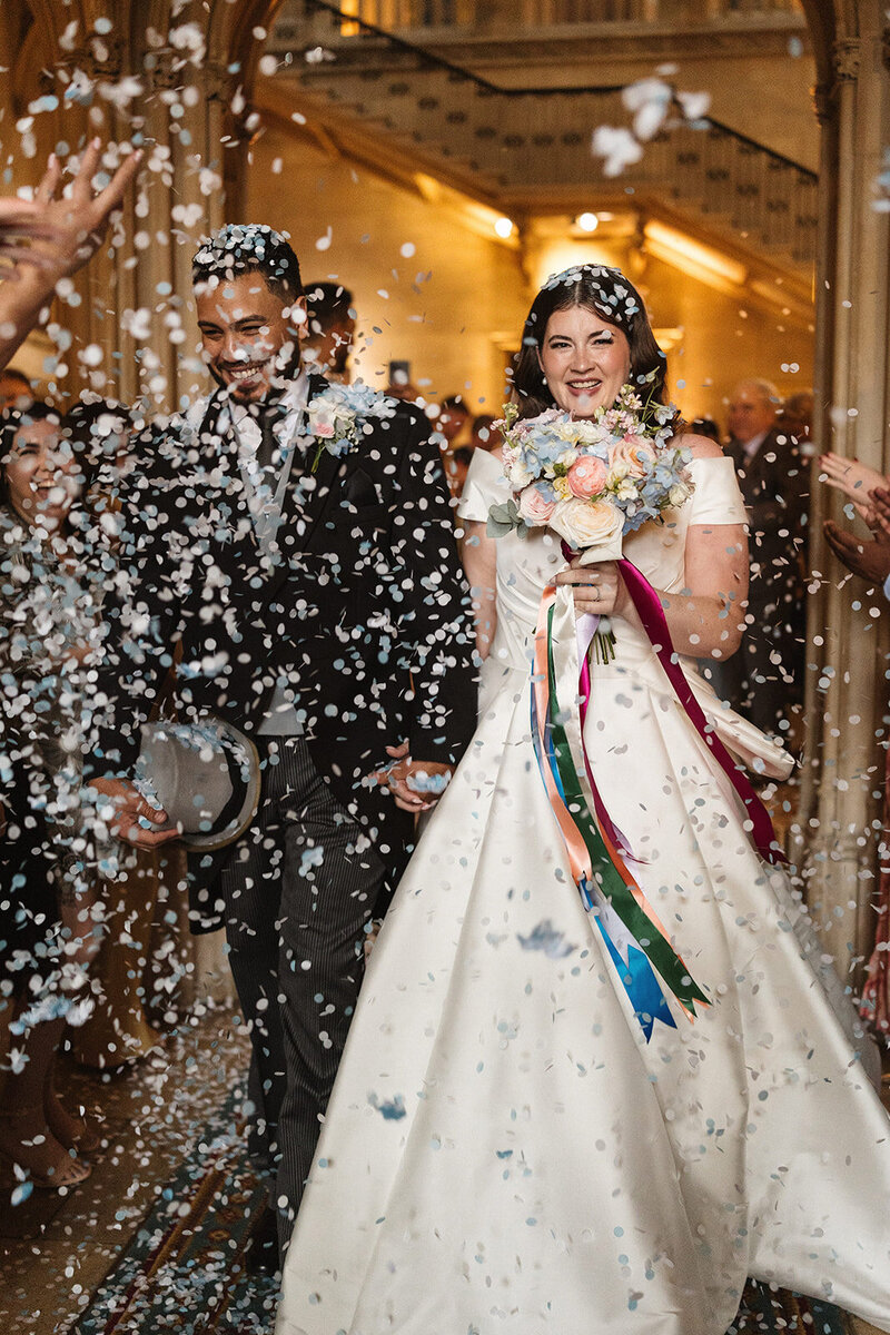 A newlywed American couple walks hand in hand through a joyful confetti line at Ashridge House, surrounded by cheering guests. The bride, glowing with happiness, holds a stunning bridal bouquet, while delicate blue and white tissue confetti flutters around them. Their beaming smiles capture the pure joy of the moment as they celebrate their love in this breathtaking setting.
