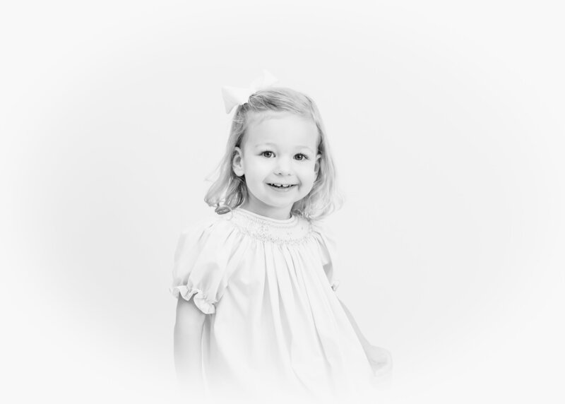 Little girl smiling in a light-colored smocked dress with a bow in her hair.