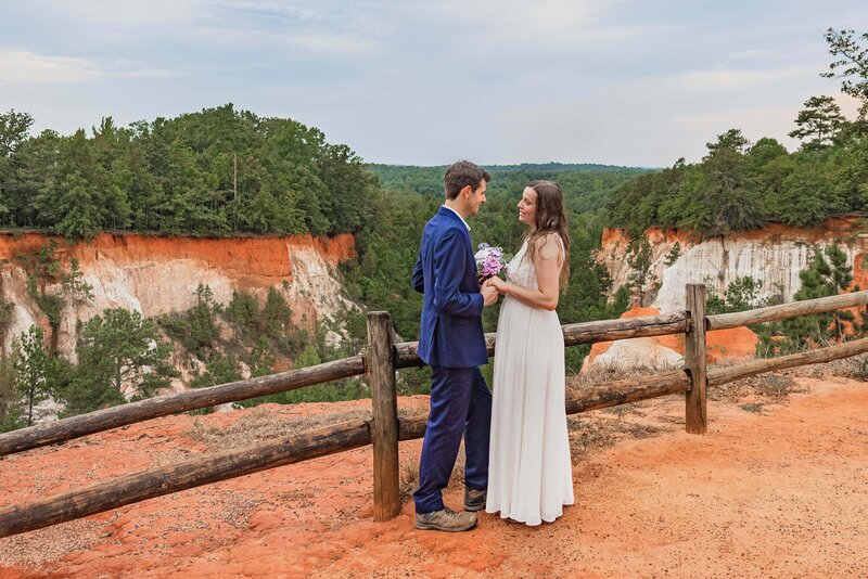 A bride and groom stand facing each other. They are reading vows and looking lovingly at each other smiling during their elopement ceremony. The bride is in a long flowing dress and the groom is in a navy blue suit. They are standing on red dirt overlooking a canyon made of red, orange and white dirt with green trees in the distance.