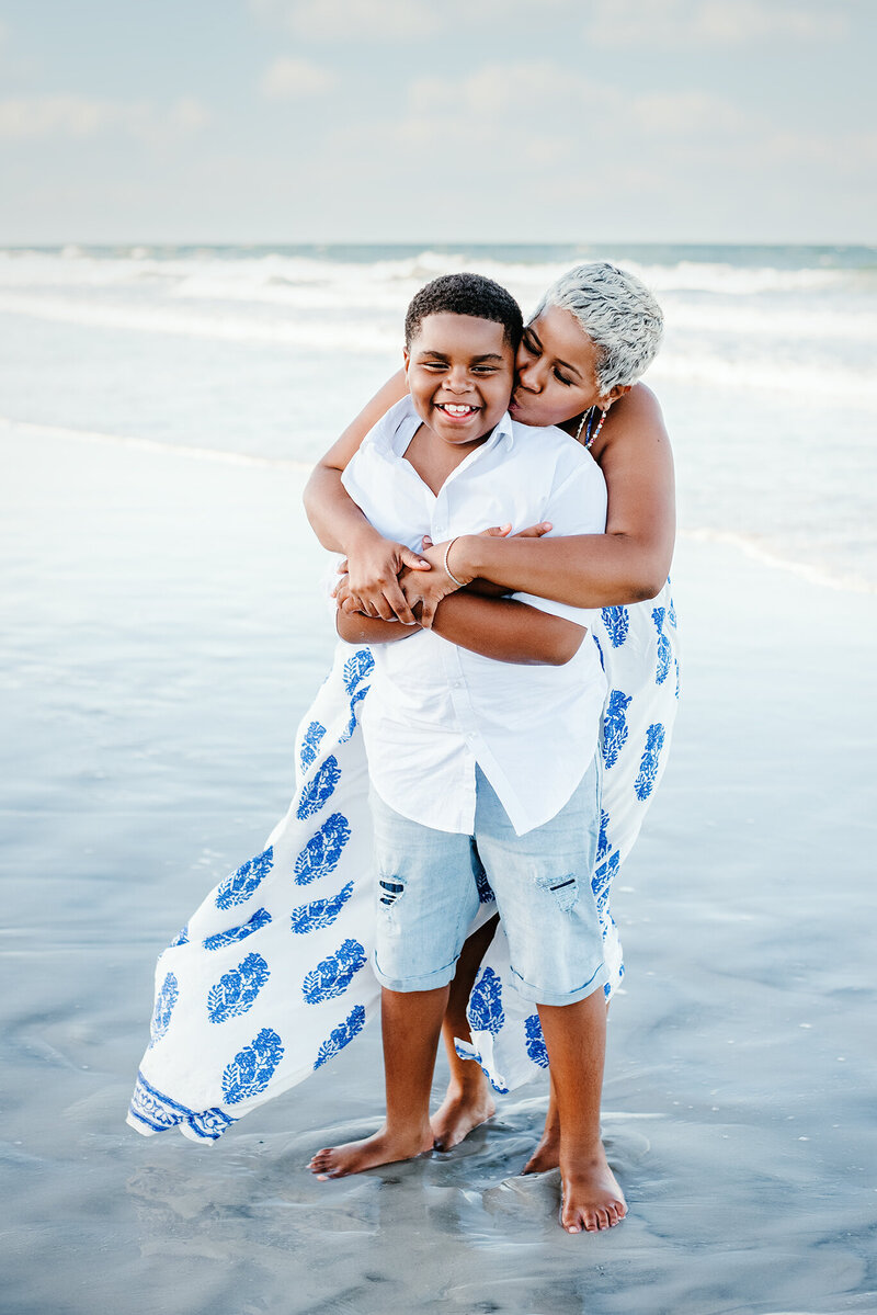Mother hugging and kissing son, dressed in blues and white, on shore of Atlantic Beach, FL.