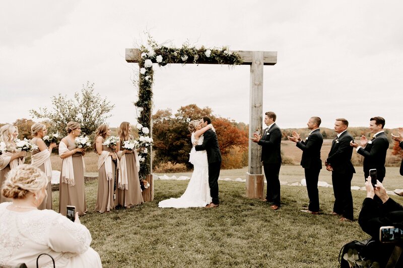 bride and groom under rustic wedding arch kissing during wedding ceremony
