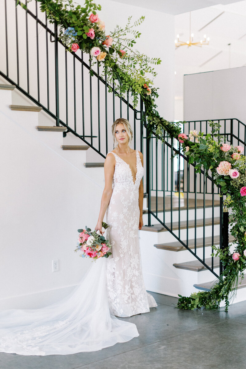 Elegant bride in a luxury gown holding a floral bouquet, standing by a staircase filled with roses at The Hutton House in Minneapolis.