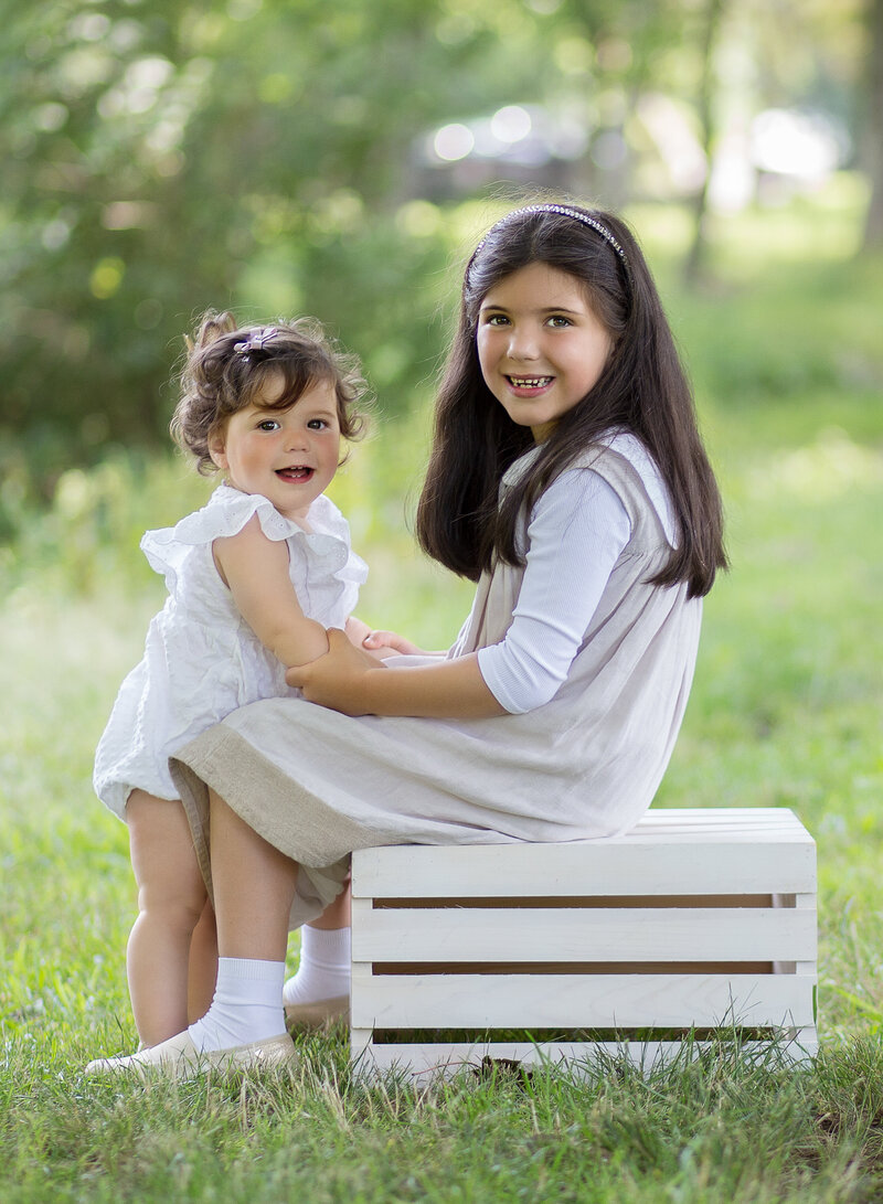 Brooklyn outdoor family photoshoot. Two sisters pose in a park. Big sister is sitting on a white crate, the little sister is standing facing her sister. Both girls are looking over their shoulder smiling. Captured by best Brooklyn, NY family photographer Chaya Bornstein.