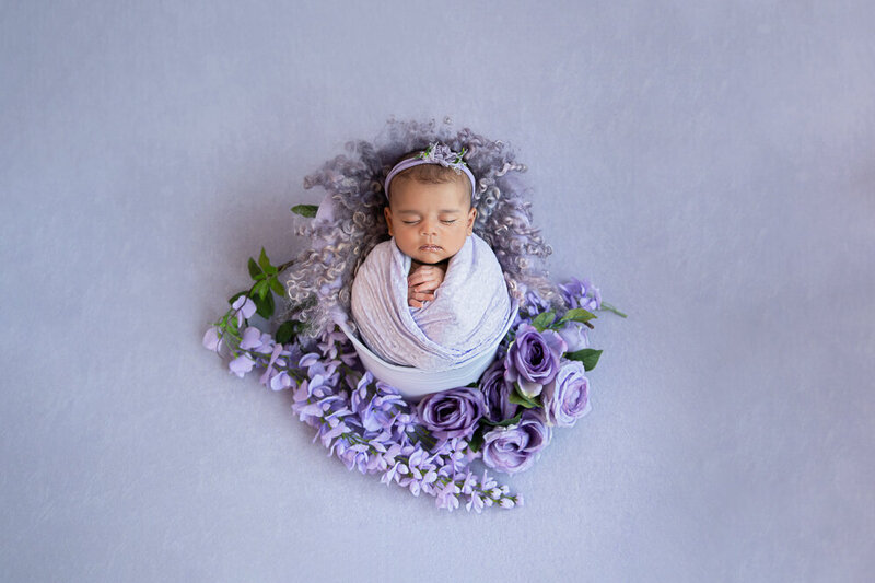 newborn girl wrapped in purple in a purple bucket with purple flowers for her newborn photography session in Burlington, Ontario