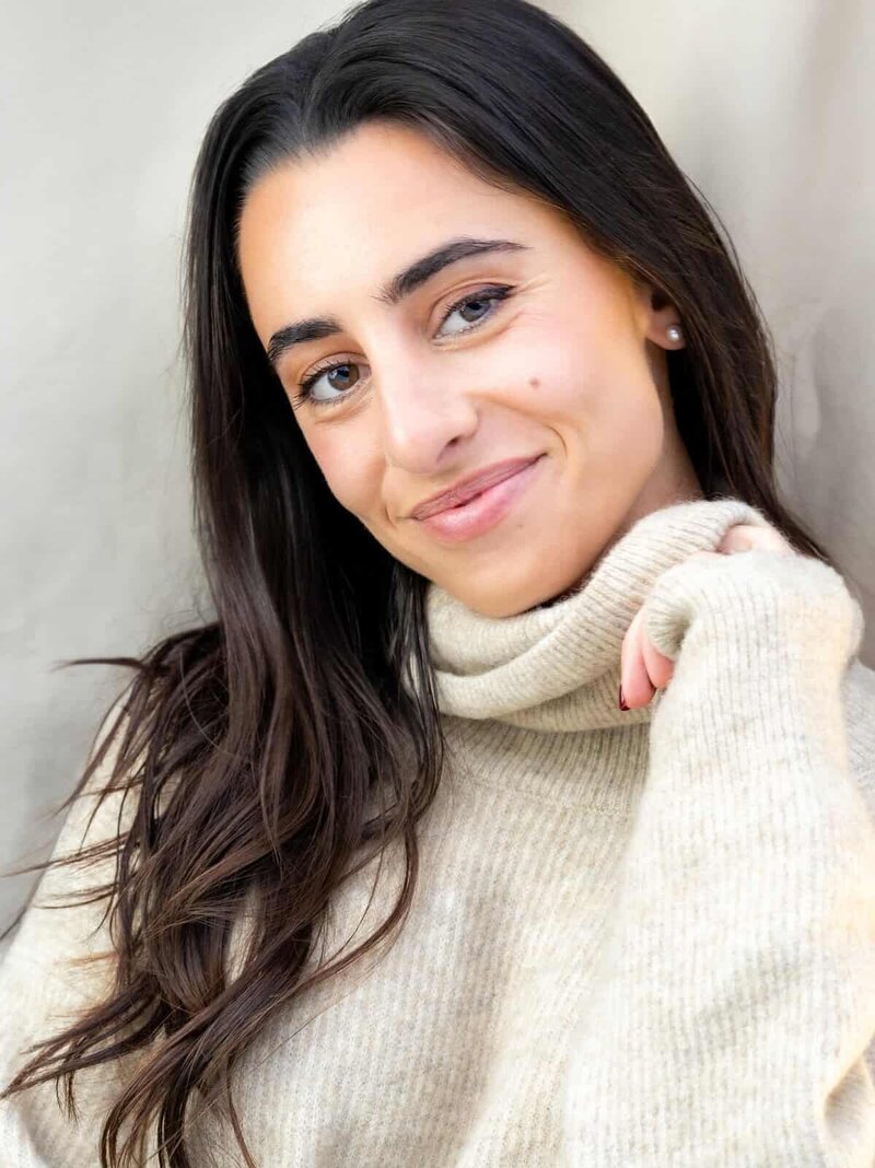 Headshot of a smiling woman with long dark hair leaning against a wall in an oversized turtleneck sweater