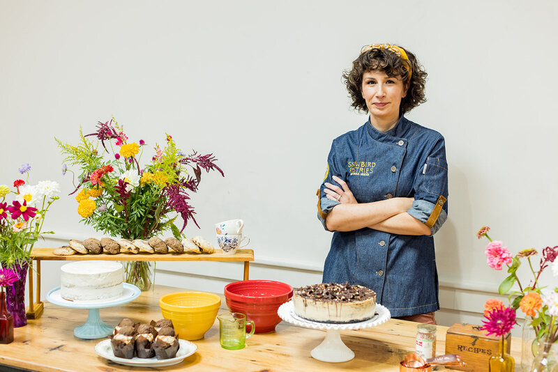 Female pastry chef in a denim jacket with embroidered branding stands confidently behind a wooden table filled with handmade desserts, fresh flowers, and vintage kitchenware. Cakes, cookies, and muffins are displayed on elegant stands, creating a warm, artisanal feel. Small business branding photography in Columbia, SC, highlighting local bakers, culinary artistry, and creative entrepreneurship.