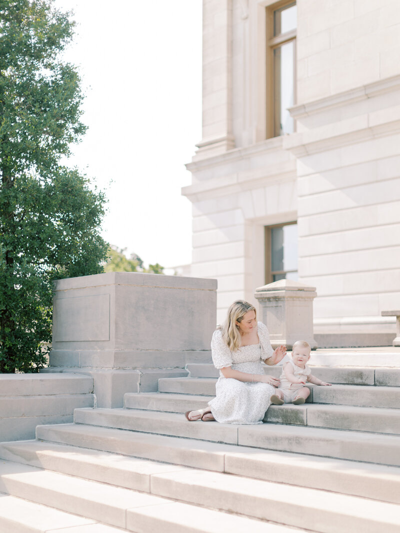 Blonde mother in neutral dress sits on Arkansas State Capitol's steps with one year old blonde son taken by Little Rock photographer Bailey Feeler