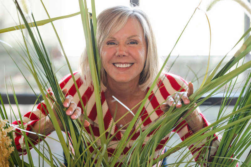 A smiling, fair-skinned woman with short, blonde hair peeks through tall green grasses or plants. She is wearing a red and white striped sweater and her hands are visible among the leaves. The background is out of focus and appears to be bright, likely an outdoor or brightly lit indoor setting.