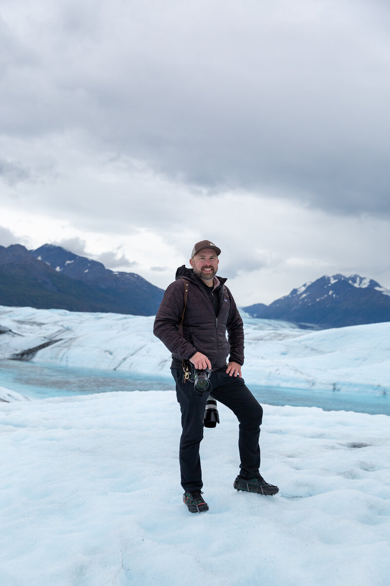 Brian, of Scenic Vows, stands on a glacier while holding a camera.