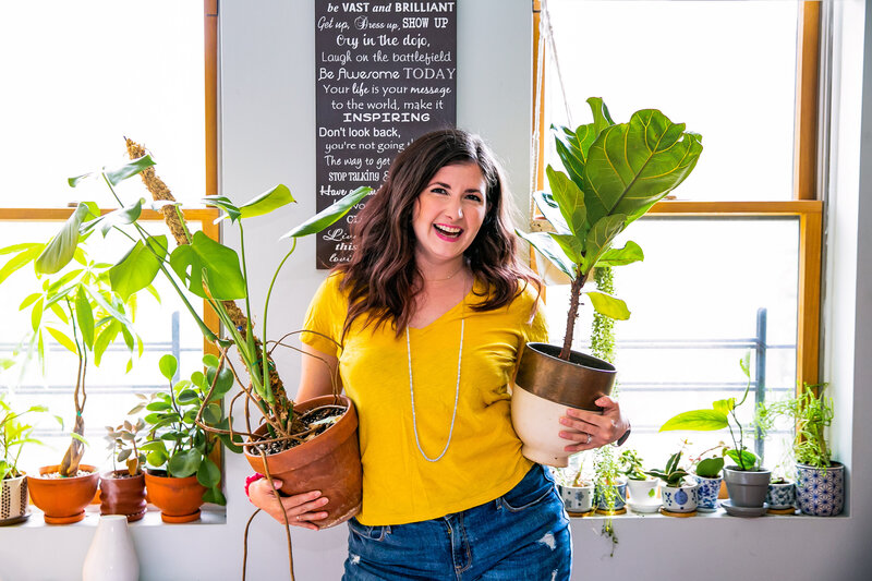 Maria smiles while holding tow large potted plants in her apartment