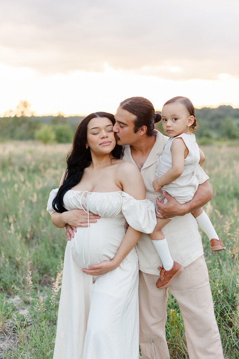 Dad holding his son and kissing the pregnant mother in a tall grass field