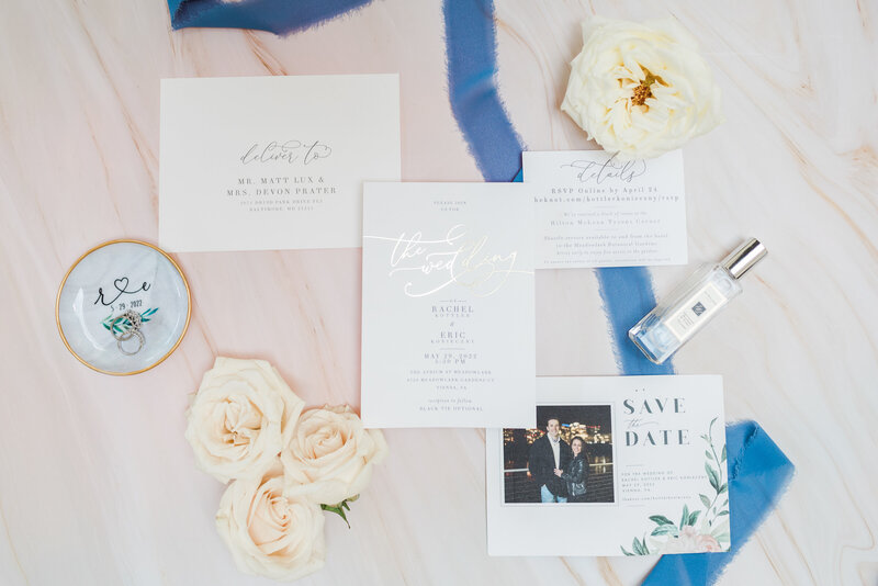 flat lay of invitation suite with a blush chiffon background and dusty blue chiffon ribbon and ivory flowers 