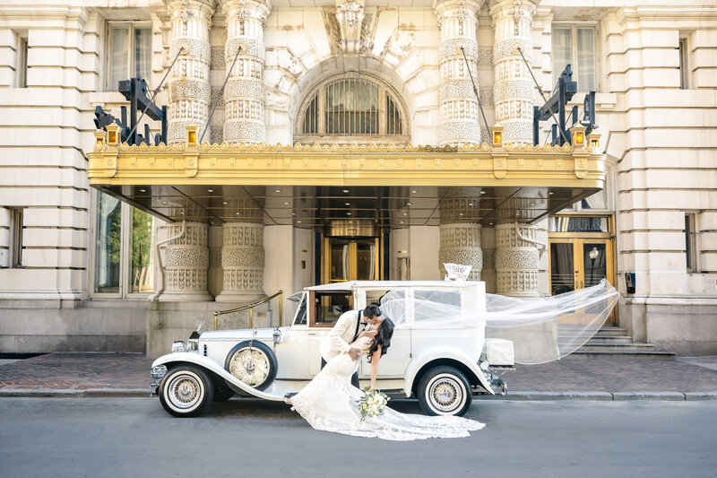 A couple shares a romantic kiss in front of a vintage white car. The bridal veil flows gracefully behind them.
