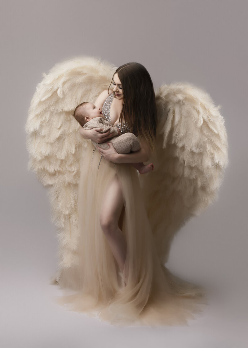 mother holding baby wearing couture feather wings in stuido setting posed for their photo shoot on grey backdrop near dupont
